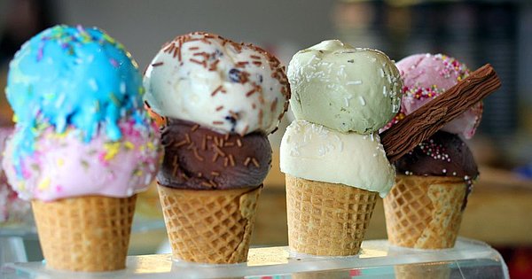 Vier Eistüten mit jeweils zwei verschiedenen Eissorten und teilweise bunten Streuseln stecken in einem Eistütenhalter.